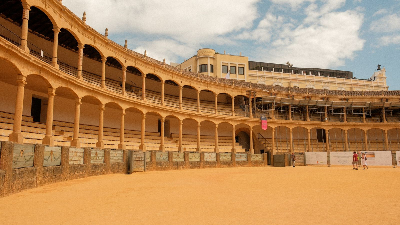 An empty bullring, with two tourists crossing the sand beneath the arcades of the stands
