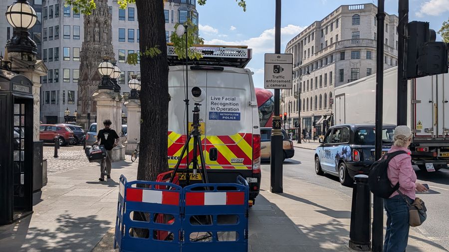 Una furgoneta de la Policía Metropolitana en una acera de Londres, con una cámara sobre un trípode y el cartel «Live Facial Recognition In Operation», mientras la gente pasa