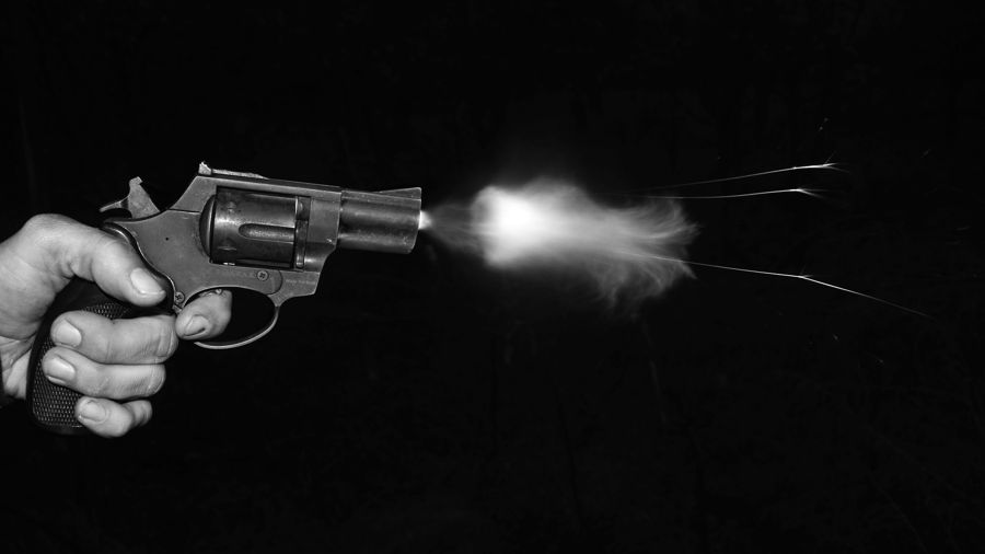 Foto en blanco y negro de una mano disparando un revólver, con el fogonazo iluminando un fondo negro