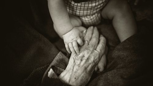 An older woman holding a baby's hand, sitting by a window