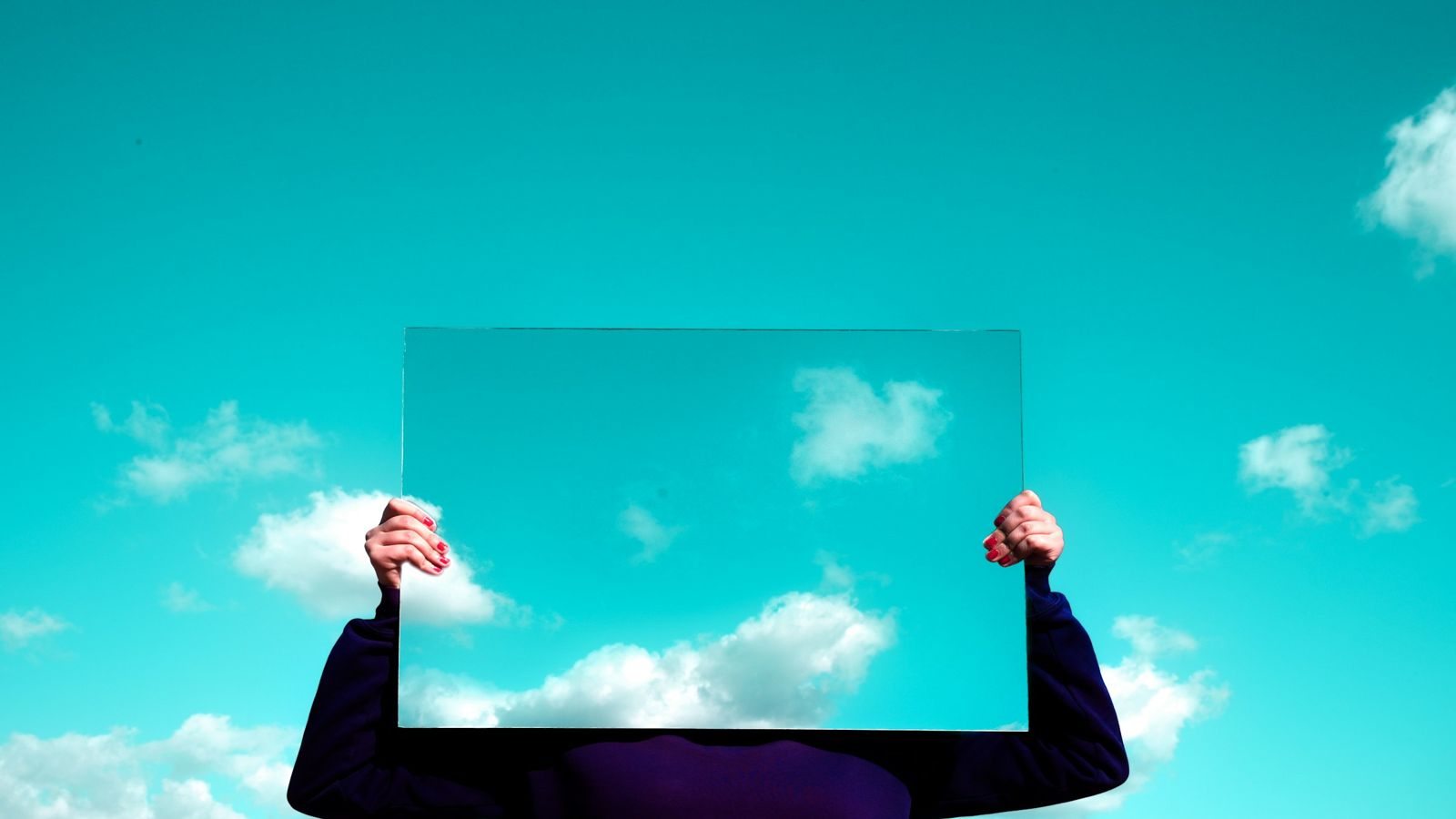 A person holds a mirror in front of their face, and the mirror shows only the sky and clouds