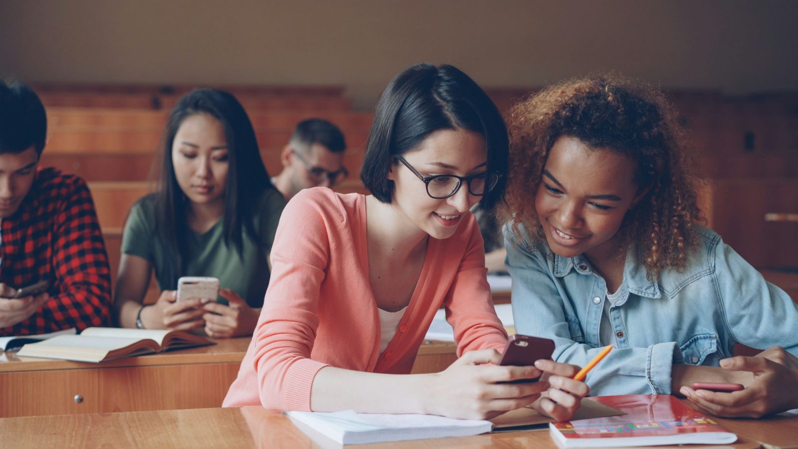 Estudiantes en un aula con gradas mirando el móvil; dos de ellas comparten uno y sonríen