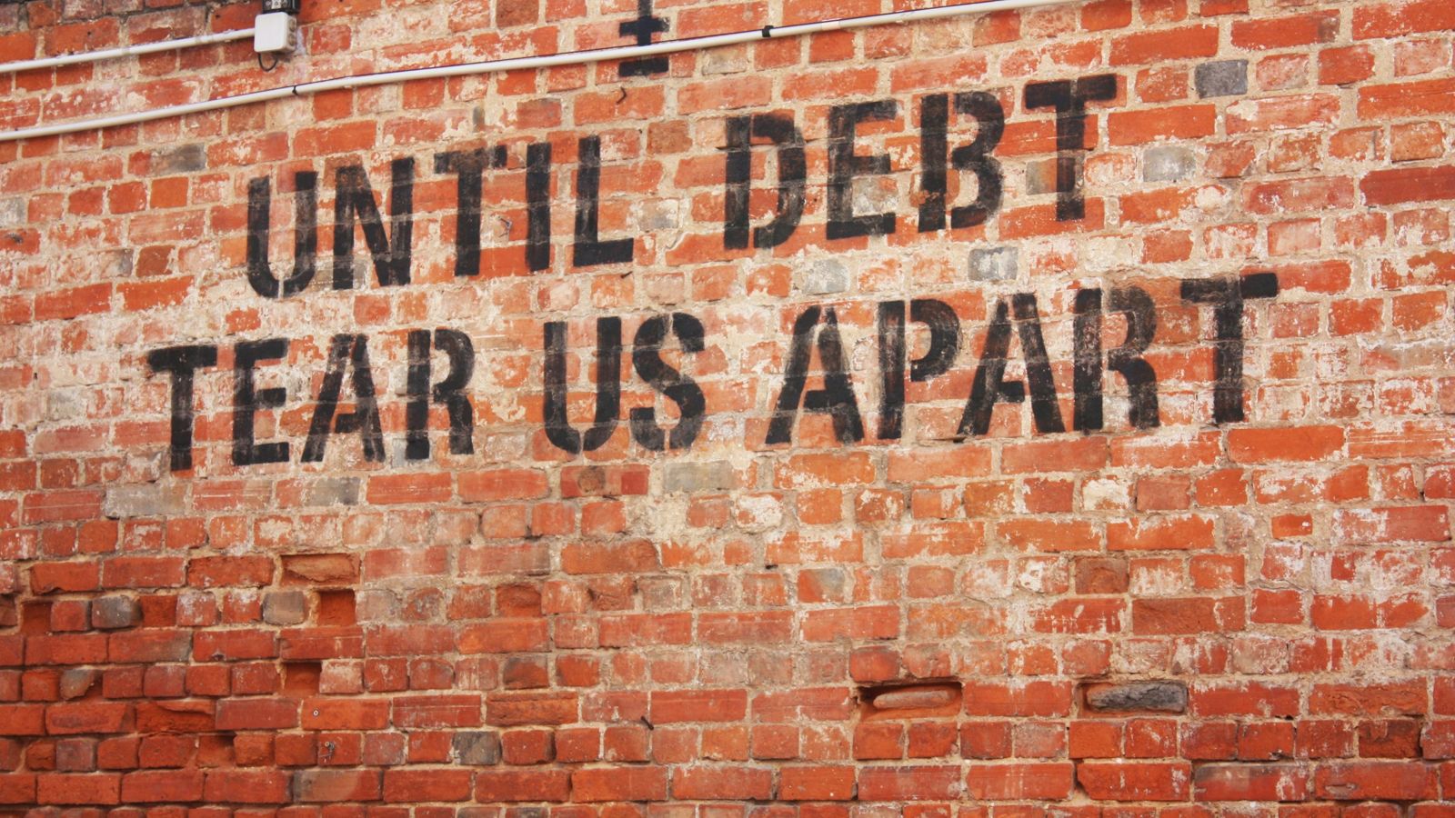 «Until debt tear us apart» —hasta que la deuda nos separe— pintado en negro sobre una pared de ladrillo, con una cámara de vigilancia encima